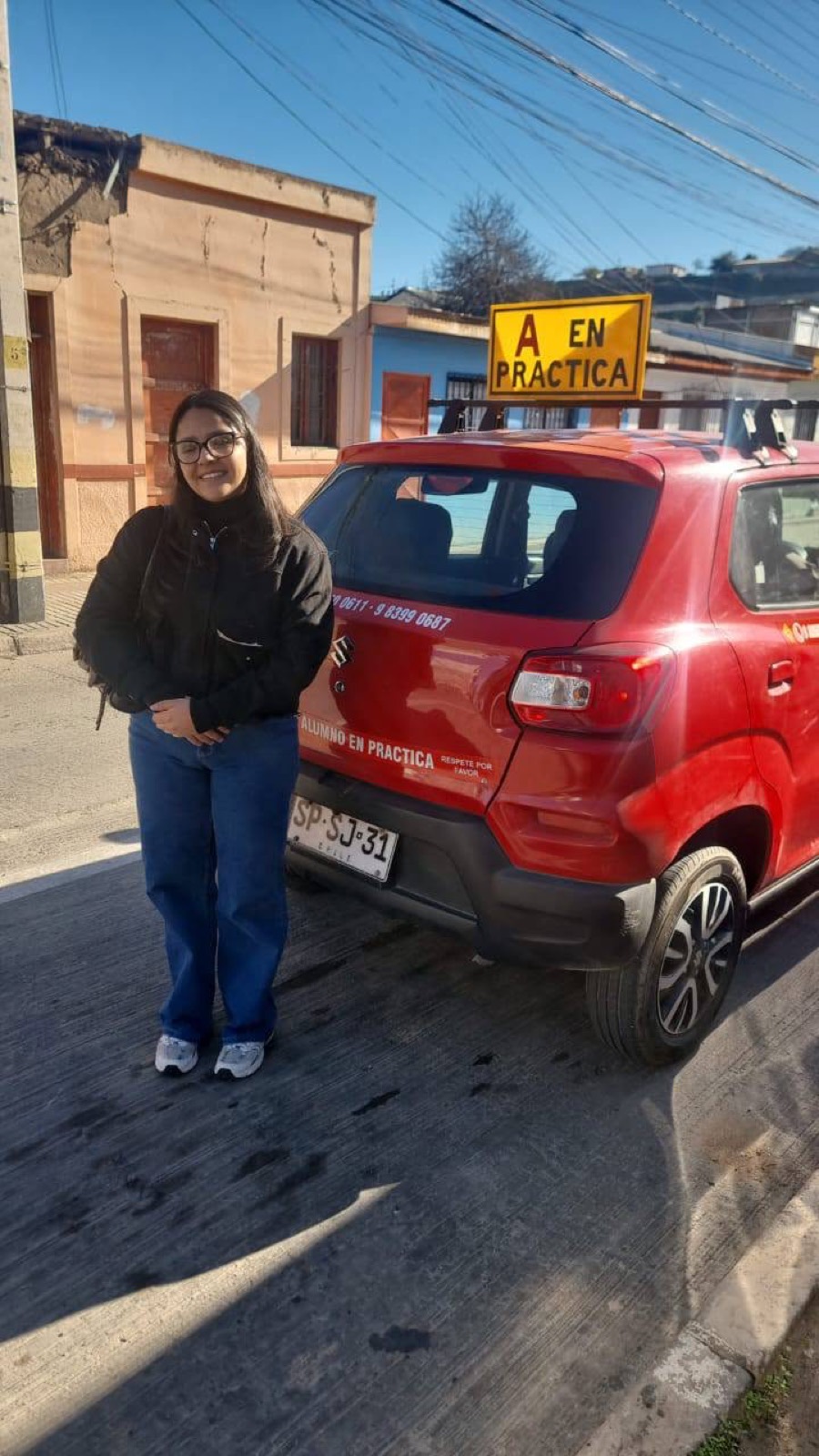 Alumna con lentes junto al auto rojo de la escuela con el cartel A en práctica, en Vallenar
